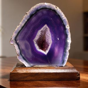 Könnte beinhalten: Eine polierte, lila Achat-Geodenscheibe, montiert auf einem rechteckigen Holzsockel. Die Geode hat ein kristallines Zentrum und ein tiefviolettes Inneres mit einem weißen und grauen äußeren Rand. Der Naturstein wird auf einem dunklen Holztisch präsentiert.