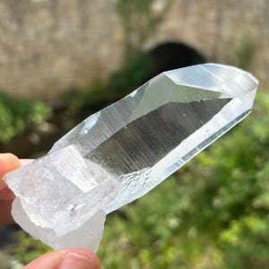Könnte beinhalten: Ein klarer, länglicher Quarzkristall mit einer scharfen, facettierten Spitze und einer sekundären, undurchsichtigen weißen Kristallbasis an einer Seite. Der Hauptkörper ist transparent und weist sichtbare Streifen und Risse auf.
