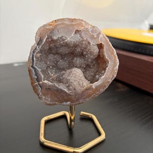 Könnte beinhalten: Eine kreisförmige Achat-Geode mit einem hohlen Zentrum, das mit kleinen, hellbraunen kristallinen Strukturen gefüllt ist. Der äußere Rand zeigt konzentrische Bänder in Beige und Weiß. Die Geode ist auf einem sechseckigen goldfarbenen Metallständer montiert.
