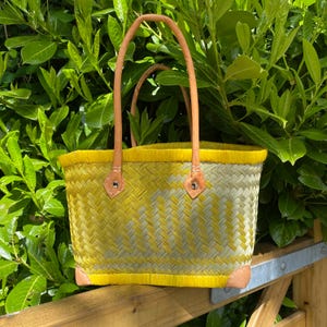 May include: A woven basket bag with a yellow and light green pattern. The bag has a yellow trim, leather handles, and leather corner reinforcements. The bag is on a wooden fence with green foliage behind it.