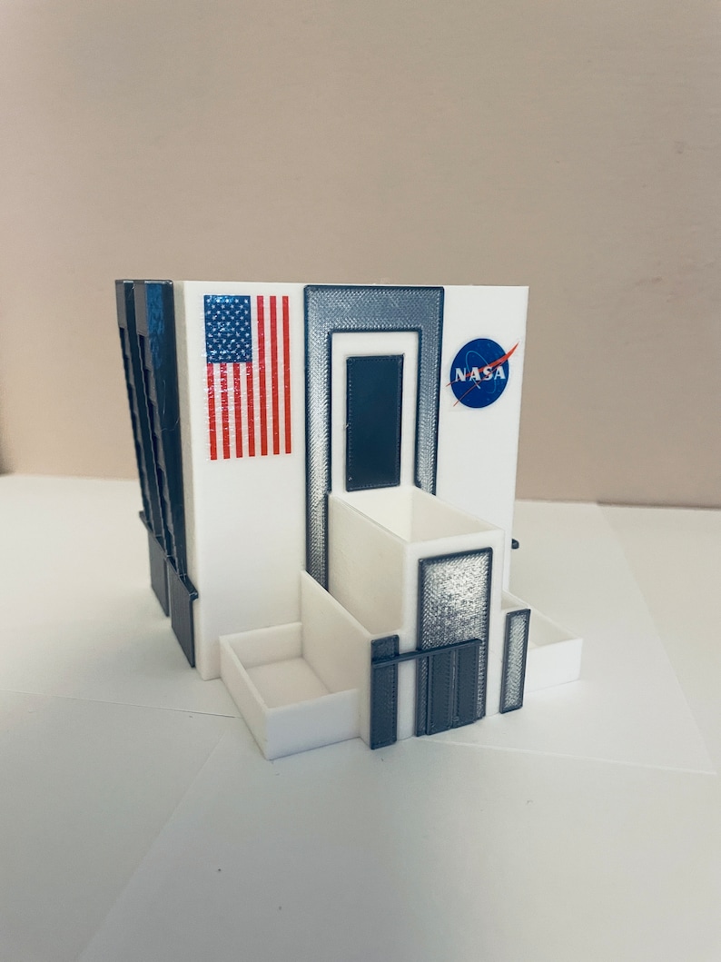K&ouml;nnte beinhalten: Ein wei&szlig;es 3D-gedrucktes Modell eines Geb&auml;udes mit einem NASA-Logo, einer amerikanischen Flagge und einer dunkelgrauen T&uuml;r. Das Geb&auml;ude hat einen kleinen Balkon und eine rechteckige &Ouml;ffnung an der Seite.