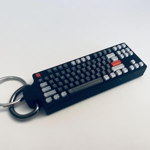 May include: A black keyboard-shaped keyring with silver key details and a red accent key. The keyring has a silver key ring attached. The product is a miniature replica of a computer keyboard.