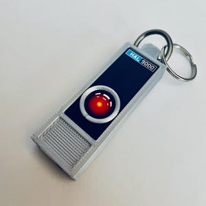 Puede incluir: Un llavero plateado con una imagen en blanco y negro de HAL 9000 de la película 2001: Una odisea del espacio. La imagen incluye el ojo rojo de HAL 9000 y el texto "HAL 9000".