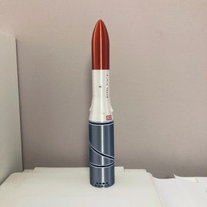 May include: A white and grey model rocket with a red nose cone. The rocket has a black and white stripe pattern and the text "BLACK ARROW" printed on the side.
