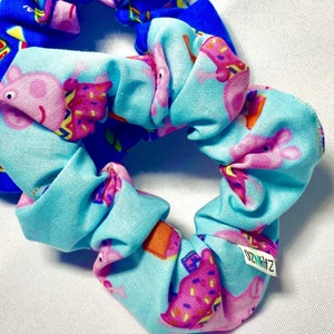 May include: A blue and light blue scrunchie with a cartoon pig design. The scrunchie has the text "ZAYHZ" on it.