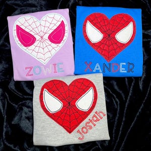 May include: Three shirts with a red heart-shaped design featuring a Spiderman mask. The shirts are in pink, blue, and gray. The names "Zowie", "Xander", and "Josiah" are embroidered on the shirts.
