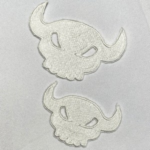 May include: Two white embroidered iron-on patches of a skull with horns. The skull has a menacing expression with large eyes and a wide mouth.
