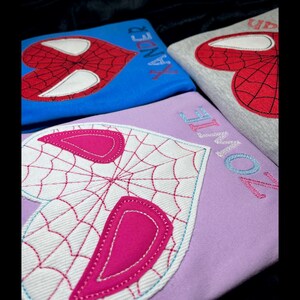 May include: Three embroidered shirts with a heart-shaped Spiderman design. The shirts are blue, grey, and purple. The names "Xander", "Zoie", and "Zoie" are embroidered on the shirts.
