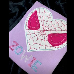 May include: A purple shirt with a white heart-shaped spider web design and the name "Ellie" embroidered in pink, blue, and white letters.