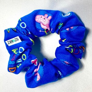 May include: Blue scrunchie with a repeating pattern of a cartoon pig and the words "Oink Oink" in a bright yellow, green, and orange font. The scrunchie has a small white tag with the text "ZAYNZ0" in green.