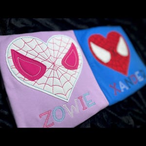 May include: Two purple and blue shirts with embroidered Spider-Man designs and the names "Zowie" and "Xander".