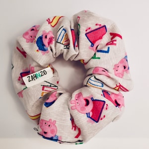 May include: A white fabric scrunchie with a pink pig cartoon character print. The scrunchie has a green label with the text "ZAYNZO" on it.
