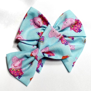 May include: A blue fabric hair bow with a Peppa Pig design. The bow is made of two large loops and a smaller loop in the center. The Peppa Pig design features Peppa Pig roller skating with a pink and purple outfit.