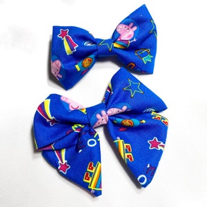 May include: Two blue fabric hair bows with a cartoon pig and star print. The bows are tied in a bow shape and have a yellow, red, and blue color scheme.