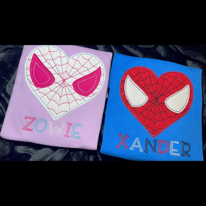 May include: Two embroidered shirts with a heart-shaped Spiderman design. The shirt on the left is purple with the name "ZOWIE" embroidered below the design. The shirt on the right is blue with the name "XANDER" embroidered below the design.
