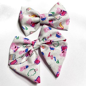 May include: Two white fabric hair bows with a pink pig cartoon print. The bows have a white background with pink, blue, and green accents. The text "PLAY" is printed on the bows.