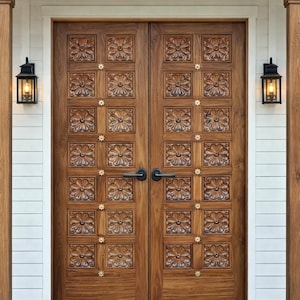 Op de afbeelding: Een paar houten deuren met ingewikkelde bloemsnijwerken. De deuren zijn warmbruin van kleur en hebben een patroon van gebeeldhouwde vierkanten. Kleine goudkleurige accenten zijn geplaatst. Zwarte lantaarns flankeren de deuren.