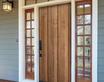 May include: A wooden front door with vertical panels, flanked by sidelights with multiple panes of glass. The door has a black handle and is set within a white-trimmed frame. The porch has a wooden floor and gray siding.