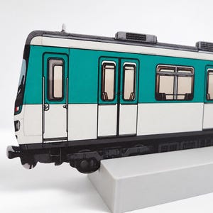 May include: A flat, two-dimensional cutout of a tube train carriage featuring a teal and white exterior colour scheme. The train carriage includes detailed windows, doors, and black undercarriage components, mounted on a solid, light grey rectangular base.