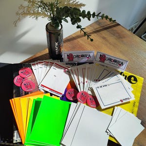 May include: A collection of stationery items, including stickers, notepads, and envelopes, arranged on a wooden surface. The items feature a cartoon character and the word "Chromatica". A spray paint can with the word "Black" is also visible.