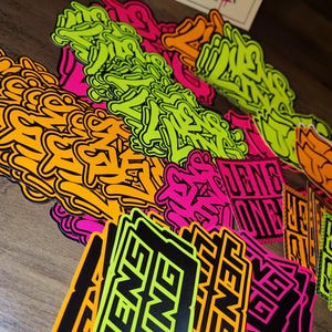 May include: A collection of colorful stickers with various designs, including the word "FENG" in black and green, black and orange, and black and pink. The stickers are on a wooden surface.