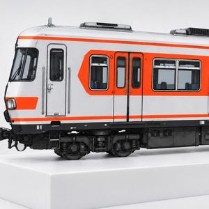 Op de afbeelding: Een modeltreinwagon met een witte buitenkant voorzien van horizontale oranje strepen, rechthoekige ramen en gedetailleerde zwarte onderstellen. De wagon heeft een voorruit, een zijdeur en een reeks passagiersramen langs de zijkant.