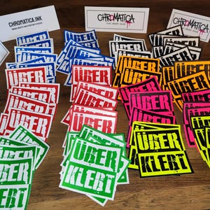 Op de afbeelding: Een verzameling kleurrijke stickers met de tekst "&uuml;ber klebt" in witte letters op een zwarte achtergrond. De stickers zijn gerangschikt in rijen en kolommen op een houten oppervlak. De stickers zijn in verschillende kleuren, waaronder rood, blauw, groen, geel en roze.