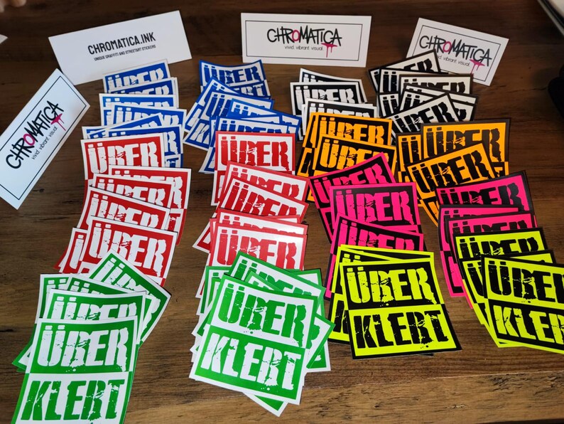 Op de afbeelding: Een verzameling kleurrijke stickers met de tekst "&uuml;ber klebt" in witte letters op een zwarte achtergrond. De stickers zijn gerangschikt in rijen en kolommen op een houten oppervlak. De stickers zijn in verschillende kleuren, waaronder rood, blauw, groen, geel en roze.