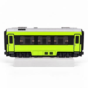 Könnte beinhalten: Ein Modelleisenbahnwagen mit einem leuchtend hellgrünen Gehäuse und einem horizontalen schwarzen Streifen in der Mitte. Das Dach ist silberfarben und das Fahrgestell ist schwarz mit sichtbaren Rädern. Der Wagen steht auf einem silbernen Sockel.