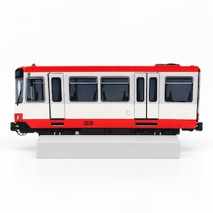 Op de afbeelding: Een modeltram met een rood en wit exterieur, zwarte raamkozijnen en donker getint glas. Het voertuig heeft een rechthoekige carrosserie met zichtbare deuren en staat op een eenvoudige, lichtgrijze rechthoekige voet voor presentatiedoeleinden.