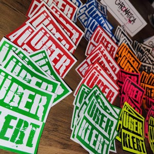 Op de afbeelding: Een verzameling kleurrijke stickers met het woord "Uber" erop gedrukt. De stickers zijn in verschillende kleuren, waaronder rood, groen, geel en zwart. De stickers zijn willekeurig gerangschikt op een houten oppervlak.