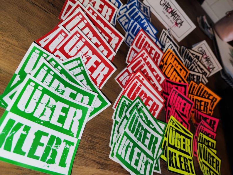 Op de afbeelding: Een verzameling kleurrijke stickers met het woord "Uber" erop gedrukt. De stickers zijn in verschillende kleuren, waaronder rood, groen, geel en zwart. De stickers zijn willekeurig gerangschikt op een houten oppervlak.