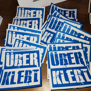 Op de afbeelding: Een stapel blauwe en witte stickers met de tekst "&uuml;berklebt" in witte letters. De stickers zijn vierkant en hebben een verweerd, grunge-ontwerp.