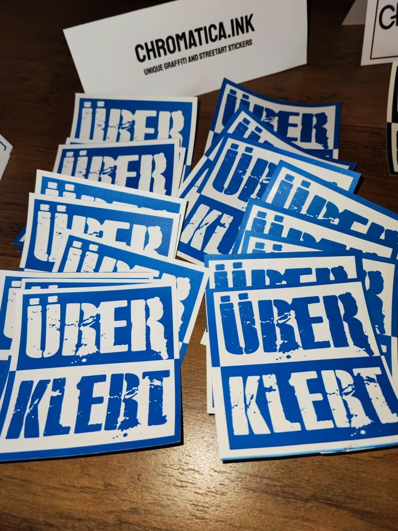 Op de afbeelding: Een stapel blauwe en witte stickers met de tekst "&uuml;berklebt" in witte letters. De stickers zijn vierkant en hebben een verweerd, grunge-ontwerp.