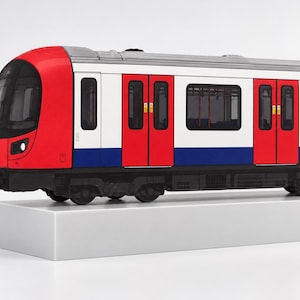 Może przedstawiać: Model wagonu kolejowego z czerwonym przodem, białymi panelami bocznymi i niebieskim dolnym pasem. Wagon posiada liczne prostokątne okna, czerwone podwójne drzwi z żółtymi etykietami ostrzegawczymi oraz czarne podwozie z widocznymi kołami.