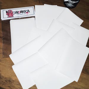 May include: Several white paper sheets, some folded, are arranged on a wooden surface. A clear plastic sleeve with a sticker that reads "CHROMATICA" and a cartoon character is visible. A black and white can of "LOOP" Pro Writing Tool is also present.