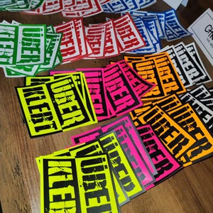 Op de afbeelding: Een verzameling kleurrijke stickers met het woord "VIBERT" in zwart gedrukt op verschillende gekleurde achtergronden, waaronder groen, rood, blauw, oranje, roze en geel.
