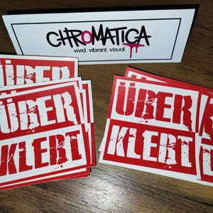 Op de afbeelding: Een stapel rode stickers met witte tekst die "&uuml;ber klerT" zegt. De stickers hebben een verweerd, grunge-achtig ontwerp.