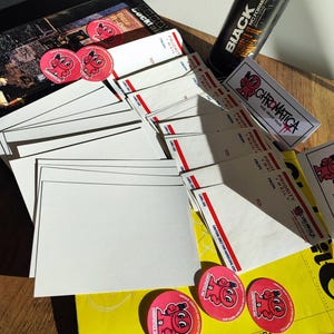 May include: An assortment of art supplies and promotional materials. Includes a black spray paint can, white envelopes with red accents, pink stickers with a cartoon character, and a black box with printed images.