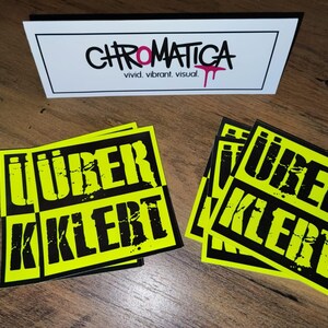 Op de afbeelding: Vier gele en zwarte stickers met de tekst "&uuml;ber kler" in zwarte letters. De stickers liggen op een houten oppervlak.