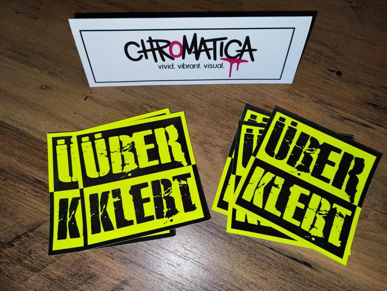 Op de afbeelding: Vier gele en zwarte stickers met de tekst "&uuml;ber kler" in zwarte letters. De stickers liggen op een houten oppervlak.