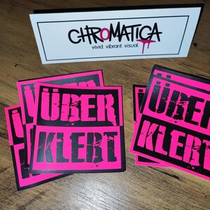 Op de afbeelding: Een stapel roze en zwarte stickers met de tekst "&uuml;ber kler" in zwarte letters. De stickers zijn vierkant en hebben een verweerde look.