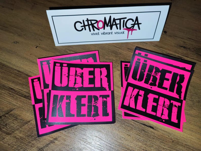 Op de afbeelding: Een stapel roze en zwarte stickers met de tekst "&uuml;ber kler" in zwarte letters. De stickers zijn vierkant en hebben een verweerde look.