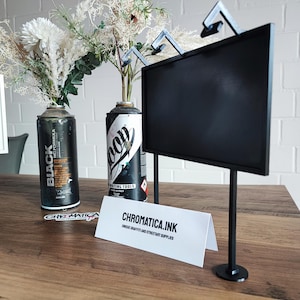 Op de afbeelding: Een zwarte displaystandaard met een rechthoekig scherm en twee steunpoten. De standaard staat op een houten oppervlak, naast twee spuitbussen die als vazen met bloemstukken worden gebruikt. Een wit bordje leest "CHROMATICA.INK".
