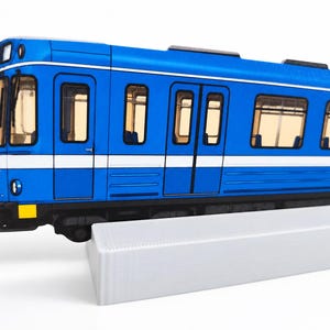 Könnte beinhalten: Ein blaues Zugwagenmodell mit einem weißen horizontalen Streifen an der Seite, schwarzen Fensterrahmen und klaren Fenstern. Der Zugwagen ist auf einem weißen rechteckigen Sockel montiert und zeigt ein kleines gelbes quadratisches Detail vorne.