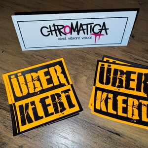 Op de afbeelding: Twee gele en zwarte stickers met de tekst "&uuml;ber klerT" in zwarte, vetgedrukte en gestileerde letters. De stickers bevinden zich op een houten oppervlak.