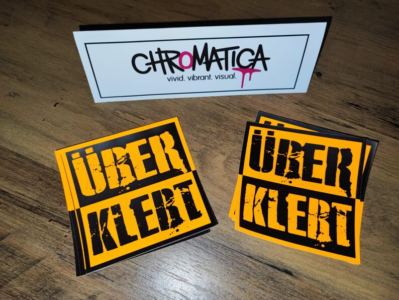 Op de afbeelding: Twee gele en zwarte stickers met de tekst "&uuml;ber klerT" in zwarte, vetgedrukte en gestileerde letters. De stickers bevinden zich op een houten oppervlak.