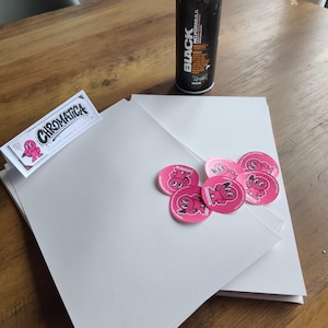 Könnte beinhalten: Ein Stapel weißer Papierbögen, ein rechteckiger Aufkleber mit dem Text CHROMATICA und einer rosa Figur sowie mehrere runde rosa Aufkleber mit derselben Figur. Eine schwarze Sprühdose mit der Aufschrift BLACK NC.FORMULA steht daneben.