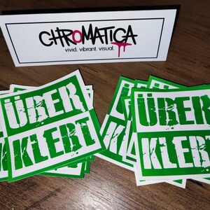 Op de afbeelding: Een stapel groene en witte stickers met de tekst "&uuml;ber kiert" erop gedrukt. De stickers hebben een verweerd lettertype in grunge-stijl.