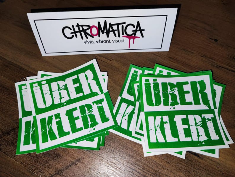 Op de afbeelding: Een stapel groene en witte stickers met de tekst "&uuml;ber kiert" erop gedrukt. De stickers hebben een verweerd lettertype in grunge-stijl.